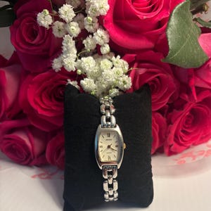 May include: A silver Seiko Luxur watch with a rectangular face and a cream-colored dial, displayed on a black velvet pillow. The watch is set against a backdrop of vibrant pink roses and delicate white baby's breath.