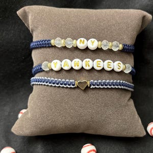Op de afbeelding: Drie handgemaakte armbanden met de woorden "NY" en "YANKEES" gespeld met witte letterkralen. De armbanden zijn marineblauw en wit, met gouden accenten en een gouden hartbedel. Honkbal-sieraden.