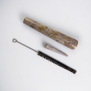 May include: A silver-coloured smoking pipe with floral carvings, a tapered mouthpiece, and a cleaning brush with black bristles. The pipe is made of metal and has a weathered appearance. The pipe is approximately 15 cm long.