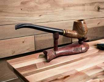 May include: A wooden pipe with a black stem resting on a wooden fish-shaped stand. The pipe is made of dark wood and has a brown bowl. The stand is made of light wood and has a carved fish design.