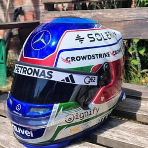 Può includere: Un casco da corsa con un design blu, rosso, bianco e verde. Presenta il logo Mercedes-Benz, la parola "PETRONAS" e il testo "CROWDSTRIKE". La visiera è scura e il casco ha la parola "Signify".