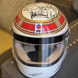 May include: A white Bell racing helmet featuring multiple black ink signatures on the top surface. The helmet includes a red and gold plaid band around the center with an "elf" logo, a clear visor, and additional signatures on the lower white section.