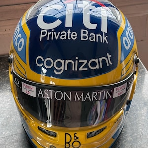 Könnte beinhalten: Ein gelber, blauer und weißer Rennhelm mit Logos von Citi Private Bank, Cognizant und Aston Martin. Der Helm hat ein Visier und den Text "B&O crypto.com icon".