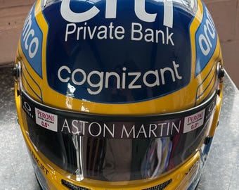 May include: A yellow, blue, and white racing helmet with the logos of Citi Private Bank, Cognizant, Aston Martin, and other sponsors. The helmet has a reflective visor and the text "B&O crypto.com icon".