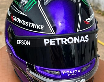 Könnte beinhalten: Ein violetter und schwarzer Rennhelm mit dem Monster Energy Logo, Crowdstrike Schriftzug und einem dunklen Visier mit Epson, Petronas, Police Eyewear und Tibco Branding. Der Helm hat ein Metallic-Finish mit weißen und grünen Akzenten.