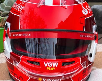 Könnte beinhalten: Ein glänzender roter Rennhelm mit weißen Akzenten und verschiedenen Sponsorenlogos wie Santander, Richard Mille, VGW Play, Shell V-Power und VistaJet. Der Helm verfügt über ein dunkel getöntes Visier, klare Belüftungsöffnungen und einen Kinnschutz.