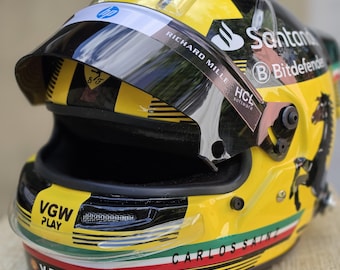 Könnte beinhalten: Ein gelb-schwarzer Rennhelm mit dem Ferrari-Logo, dem Namen Carlos Sainz und verschiedenen Sponsorenaufklebern wie HP, Santander, Bitdefender, VGW Play, V-Power und VistaJet. Der Helm hat ein dunkel getöntes Visier und italienische Flaggenstreifen.
