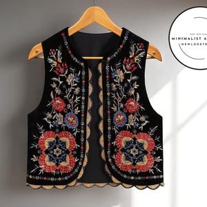 Könnte beinhalten: Schwarze Weste mit farbenfroher Blumenstickerei. Die Weste hat rote, blaue und goldene Blumendesigns. Die ärmellose Weste hat einen gewellten Rand und hängt an einem Holzbügel.