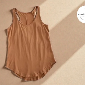 May include: A tan racerback vest with a curved hem. The top is made of a soft material and is displayed on a neutral background. A white circular logo with text is in the upper right corner.