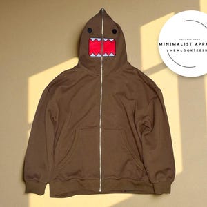 Peut inclure: Sweat à capuche marron avec un motif de visage Domo-kun sur la capuche. La capuche présente des yeux noirs, une bouche rouge et des dents blanches. Le sweat à capuche a une fermeture éclair argentée et une poche avant. Le texte "MINIMALIST APPAREL" et "NEWLOOKTEESBYOMRI" est dans un cercle.