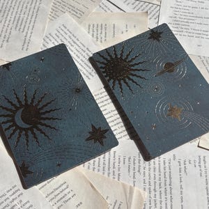 May include: Two rectangular, celestial-themed cards with a dark teal background. One card features a sun and crescent moon design, while the other has a sun, planets, and shooting stars. The cards are placed on a background of aged book pages.