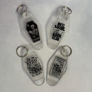 May include: Four clear, glittery keychains with silver key rings. Each keychain has a different black graphic and text design. The designs include a skeleton reading in a coffin, a book with the words "Anti Social Book Club", and more.