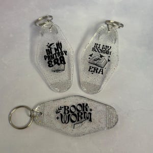 May include: Three clear, glitter-filled keychains with silver key rings. Two keychains have an oval shape and the text "IN MY FANTASY ERA" and "IN MY BOOKISH ERA". The third keychain is rectangular and reads "BOOK WORM".