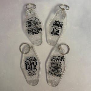 May include: Four clear, glittery keychains with silver key rings. Each keychain has a different black graphic and text: "A book a day keeps the mental breakdown away", "Happiest when I'm reading", "Books are my therapy", and "Books make me feel things."