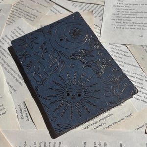 May include: A dark blue rectangular journal with an embossed celestial design featuring a sun, moon, stars, and floral elements. The journal is resting on a background of open book pages.