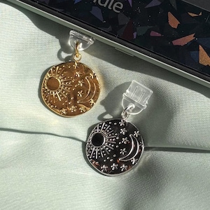 May include: Two celestial-themed earrings. One is gold-toned, the other silver-toned. Each earring features a sun, moon, and star design. The earrings are round and dangle from clear plastic earring backs. They rest on a light green surface.