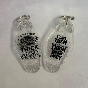 May include: Two clear acrylic keychains with silver key rings. The keychains have glitter and black text that reads "I LIKE THEM THICK & SPICY" and "I LIKE THEM THICK AND SPICY". One keychain has a book and rose graphic.