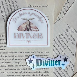 May include: Two stickers on a book page. One is a white rectangle with a moth illustration and the text "It's been a privilege DIVINER". The other is a holographic sticker with the word "Diviner".