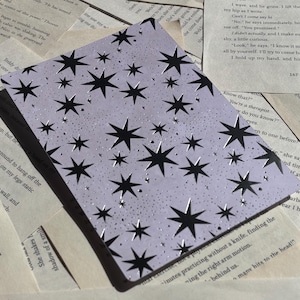 May include: A light purple notebook with a black star pattern. The stars vary in size and are scattered across the cover. The notebook is lying on top of pages from a book.