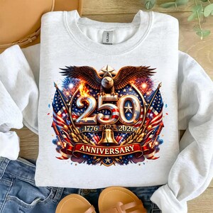 May include: White crew neck sweatshirt featuring a patriotic design. The graphic includes an eagle, American flags, the number 250, and the years 1776 and 2026, with the word "Anniversary" on a banner. The design incorporates red, white, blue, and gold colors.