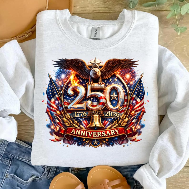 May include: White crew neck sweatshirt featuring a patriotic design. The graphic includes an eagle, American flags, the number 250, and the years 1776 and 2026, with the word "Anniversary" on a banner. The design incorporates red, white, blue, and gold colors.
