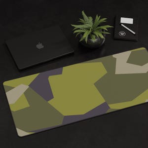 May include: A green, brown, and purple camouflage desk mat with a laptop computer, a potted plant, and a notebook on a black surface.
