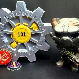 May include: A silver and yellow Vault-Tec themed display piece with the number 101, a Fallout character figurine, and two bottle caps. The figurine is holding a weapon.