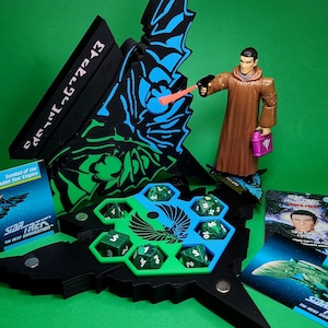 May include: A Star Trek: The Next Generation board game set with a figure, cards, and dice. The game features a black and green hexagonal dice holder, a figure in a brown robe, and cards with images of characters and ships. The background is green.