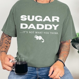 May include: Olive green t-shirt with the words "SUGAR DADDY" in large white letters. Below the text is the phrase "...IT'S NOT WHAT YOU THINK." and a white cartoon animal. The person is wearing blue jeans and holding a clear glass mug.