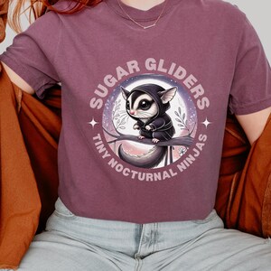 May include: A purple t-shirt with a graphic of a sugar glider wearing a black hooded cloak. The graphic is surrounded by a circle with the text "Sugar Gliders Tiny Nocturnal Ninjas".