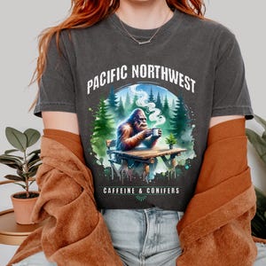 Puede incluir: Una camiseta gris oscuro con un gráfico de un Bigfoot sentado en una mesa en un bosque con una taza de café. El texto "Pacific Northwest Caffeine & Conifers" está impreso en la camiseta.