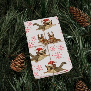 May include: A wrapped gift with a festive Christmas theme. The white wrapping paper features illustrations of sugar gliders wearing Santa hats and reindeer antlers, along with red snowflakes. The gift is tied with twine and set against a backdrop of pine needles and pine cones.