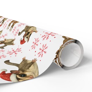 May include: White wrapping paper featuring a repeating pattern of sugar gliders wearing Santa hats and red snowflakes. The paper is rolled up, showing the design on the front.