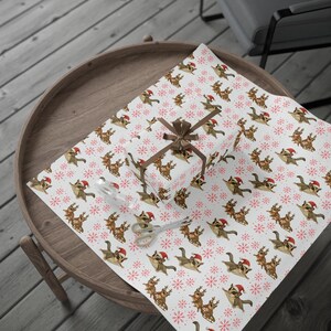 May include: A wrapped gift on a wooden table, covered in white wrapping paper with a repeating pattern of squirrels wearing Santa hats and reindeer. The gift is tied with a brown ribbon and bow. A pair of scissors is on the wrapping paper.