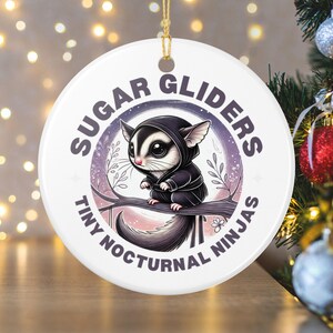 Op de afbeelding: Witte keramische kerstversiering met een ontwerp van een suikereekhoorn. De suikereekhoorn draagt een zwarte hoodie en zit op een tak. De tekst op de versiering luidt "Sugar Gliders Tiny Nocturnal Ninjas."
