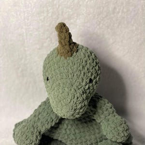 May include: A green crocheted dinosaur plush toy with a brown horn. The dinosaur is sitting with its legs crossed and has a friendly expression.