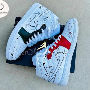 May include: Pair of white high-top sneakers with black paint splatter details. One shoe features a red accent, the other a green accent. The Nike swoosh is visible on both shoes. The shoes are on a black box.