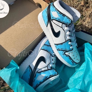 May include: A pair of high-top sneakers in a box. The shoes are primarily white and light blue, with black accents and a cartoon-like outline. The box is brown, and the shoes rest on blue tissue paper.