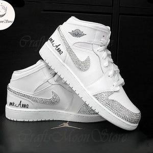 May include: White high-top sneakers with crystal embellishments on the toe, Nike swoosh, and heel. The text "mr. Anna" is written on the side of the shoe. The sneakers have white laces and a white sole.