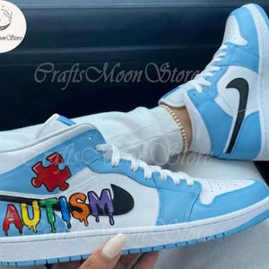 May include: Light blue and white high-top sneakers with black swoosh logos. The word "AUTISM" is written in colorful letters on the side of the shoe, along with a puzzle piece graphic. The sneakers have white laces and soles.