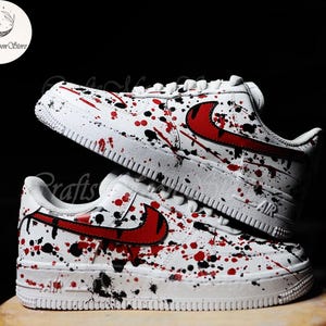 Pode incluir: Tênis brancos com design de respingos de tinta vermelha e preta. Os tênis apresentam um logotipo vermelho e cadarços brancos. Os tênis são exibidos em uma superfície marrom clara sobre um fundo preto.