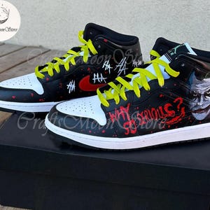 May include: Pair of custom black and white high-top sneakers with yellow laces. The sneakers feature red accents, graffiti-style text, and a Joker face design. The shoes are displayed on top of a black box.