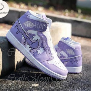May include: A pair of high-top sneakers in white and lavender. The sneakers feature a puzzle piece design with a dripping paint effect. The Nike swoosh is visible on the side. The text "Crafts Moon Store" is visible.