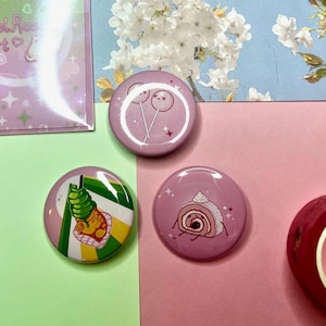 Puede incluir: Colección de impresiones artísticas y botones rosas y verdes. Los botones presentan ilustraciones de dulces. Las impresiones y botones tienen el texto "MelRoses Art". El fondo es una mezcla de rosa, verde y azul.