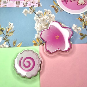 May include: Two clear acrylic charms: a pink flower and a white and pink swirl. The flower charm has a pink gradient and yellow centre. The swirl charm is white with a pink spiral. The background is blue, pink, and green.