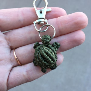 May include: A green turtle keychain charm, handmade from knotted cord. It's attached to a silver-coloured metal clasp and ring. The keychain is held in a hand against a blurred background.