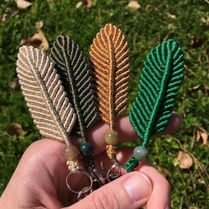 May include: Four handmade macrame feather keychains with green, brown, and beige cords and green stone beads.