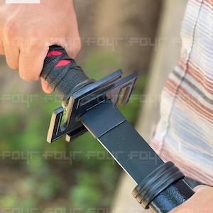 Pode incluir: Uma espada katana preta com uma empunhadura preta envolta em tecido preto com detalhes vermelhos. A espada tem uma guarda preta e uma lâmina preta. A imagem mostra a espada sendo segurada.