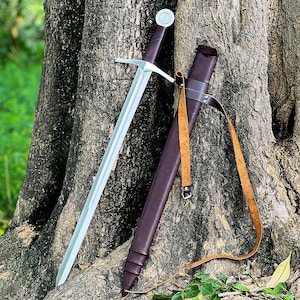 May include: A silver sword with a brown leather-wrapped handle and a matching sheath. The sword is leaning against a tree trunk. The sheath has a tan leather strap. The sword has a cross-shaped hilt.
