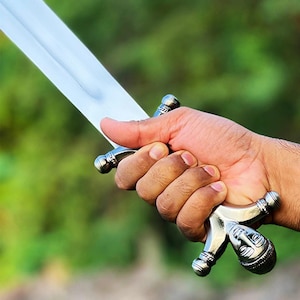 May include: A silver sword with a polished blade is held in a hand. The hilt features a detailed design with a figure's face. The background is a blurred green, suggesting an outdoor setting. The sword's blade is angled, catching the light.